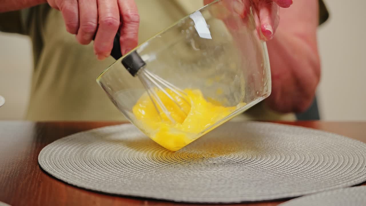 European pensioner whisks eggs in glass bowl slowly with care in kitchen light