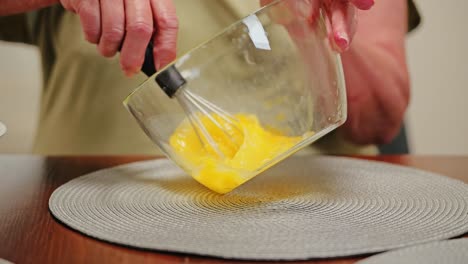 european pensioner whisks eggs in glass bowl slowly with care in kitchen light