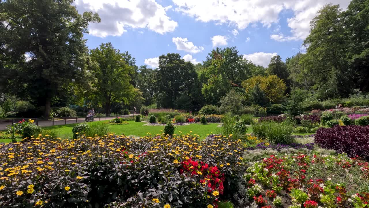 Geniş açılı gündüz kaydırma, parlak doğal güneş ışığı altında peyzajlı bir Berlin parkında canlı çiçek tarhlarını, yemyeşil bitki örtüsünü ve bir heykeli ortaya çıkarıyor.