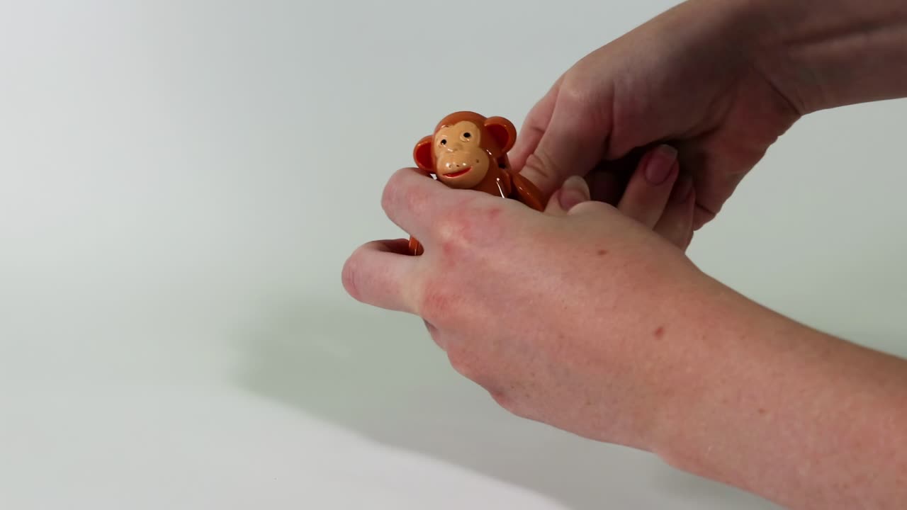 A toy monkey is manipulated by hands on a white surface, showcasing playful interaction and positioning.