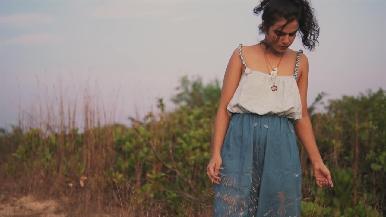mujer joven con cabello negro se para cerca de la playa y toca hierbas con la mano, chica pensativa soñadora