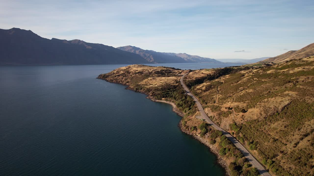 Coastal Road Along Golden Hills On Shore Of Lake Hawea In Otago Region, New Zealand. Mountain Range In Background. aerial pullback shot