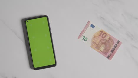 Disparo-De-Moneda-Superior-De-La-Mano-Poniendo-Un-Billete-De-10-Euros-Junto-A-Un-Teléfono-Móvil-De-Pantalla-Verde