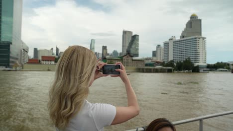 una turista tomando una foto con su teléfono y disfrutando de un paseo en barco a lo largo del río chao phraya en bangkok, tailandia
