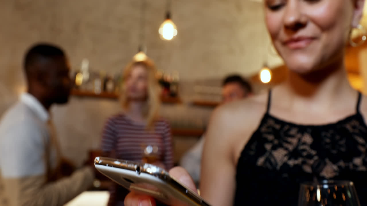una joven sonriente usando su móvil mientras sus amigos interactúan en segundo plano en el bar 4k 4k