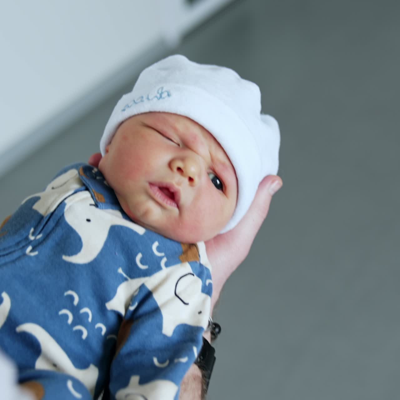 Cute dark-eyed newborn boy in blue romper and white cap. Few hours old child in dad's hands