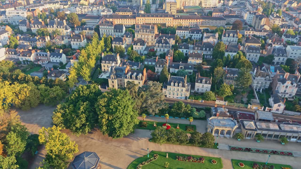 Drone shot moving backward above Thabor Park in Rennes, showing houses, trees, and the city under warm sunrise light