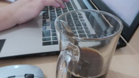 close-up of hands typing on a laptop with a glass mug of coffee and a mouse nearby.