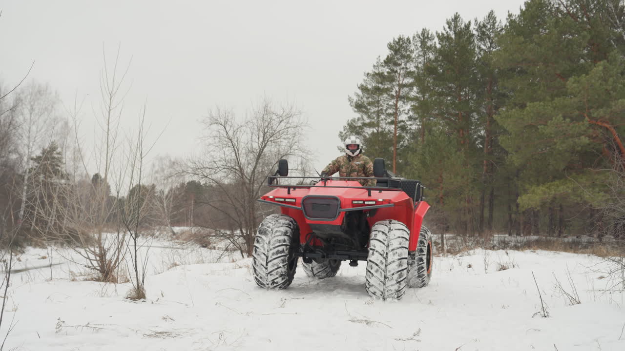 Un pilota su quad rosso in una foresta innevata. Una vista ravvicinata mostra il conducente, con casco bianco e giacca mimetica, mentre sterza nella neve fresca. Pneumatici aggressivi sollevano la neve, si sente il rombo del motore, sotto un cielo coperto e teso e pini.