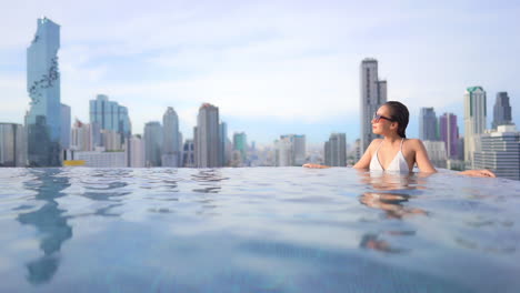 Mujer-Exótica-En-La-Piscina-Infinita-En-La-Azotea-Con-Impresionantes-Vistas-De-La-Ciudad-Moderna-En-El-Fondo,-Cuadro-Completo