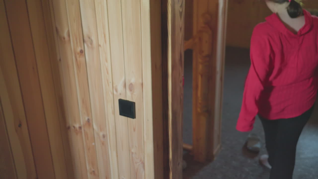 woman with ponytail in red shirt gets up from bed in cozy wooden room walks across floor and steps out of room after putting on slipper with soft lighting and casual morning indoor atmosphere