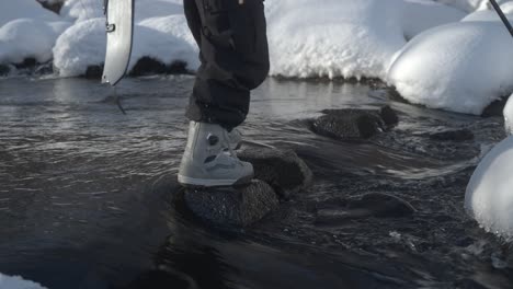 A-person-steps-on-rocks-crossing-a-snowy-stream-in-Hokkaido,-Japan,-carrying-a-splitboard