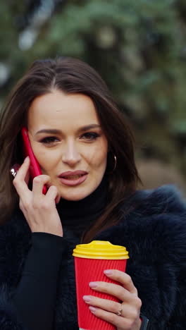girl in fur coat with a phone and coffee outdoors. portrait of a young woman talking the mobile phone and drinking hot beverage in the park. slow motion. vertical video