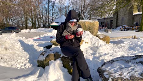 woman sits on the snow holding a cellphone and a cup on a sunny day