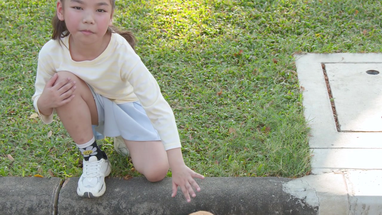 Seorang gadis muda Thailand berjongkok di taman sambil dengan lembut mengelus kucing oranye.