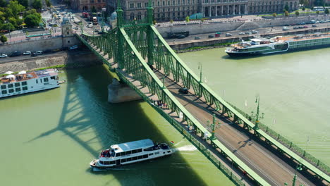 tourist boat sails under liberty bridge, aerial pull back revealing budapest