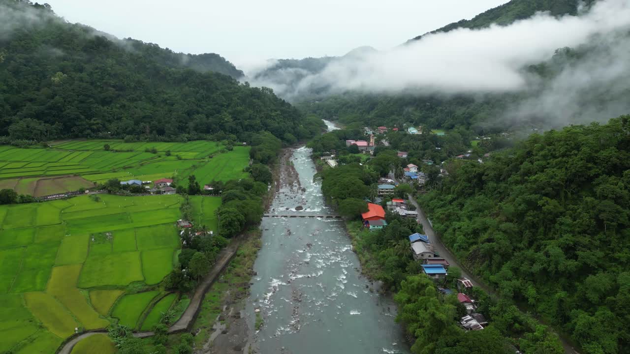 Wide aerial view of a lush Philippine valley with rice paddies, village rooftops, and a winding river framed by misty forested hills in a serene tropical landscape