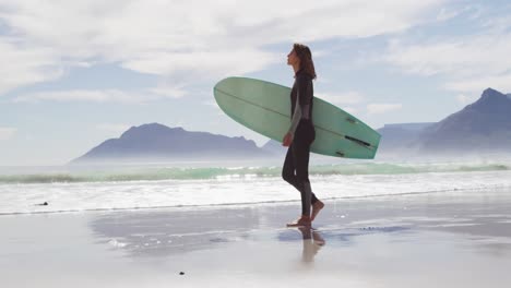 Happy-mixed-race-woman-walking-along-beach-by-the-sea-carrying-surfboard