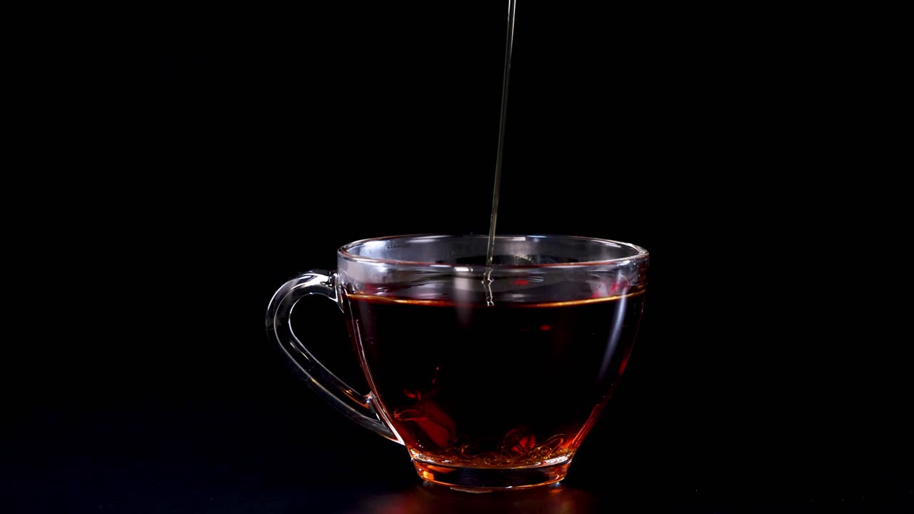 Honey streams into a glass cup of tea, creating a soothing visual against a dark backdrop