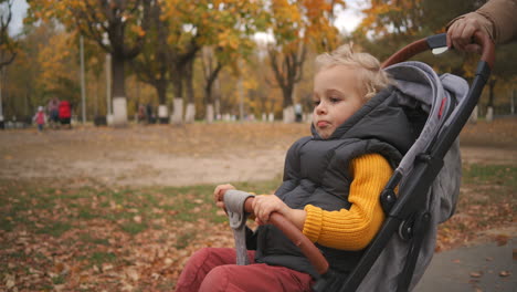 portrait of little child in stroller mother is rolling his in park area in city at autumn day rest and walk at weekend