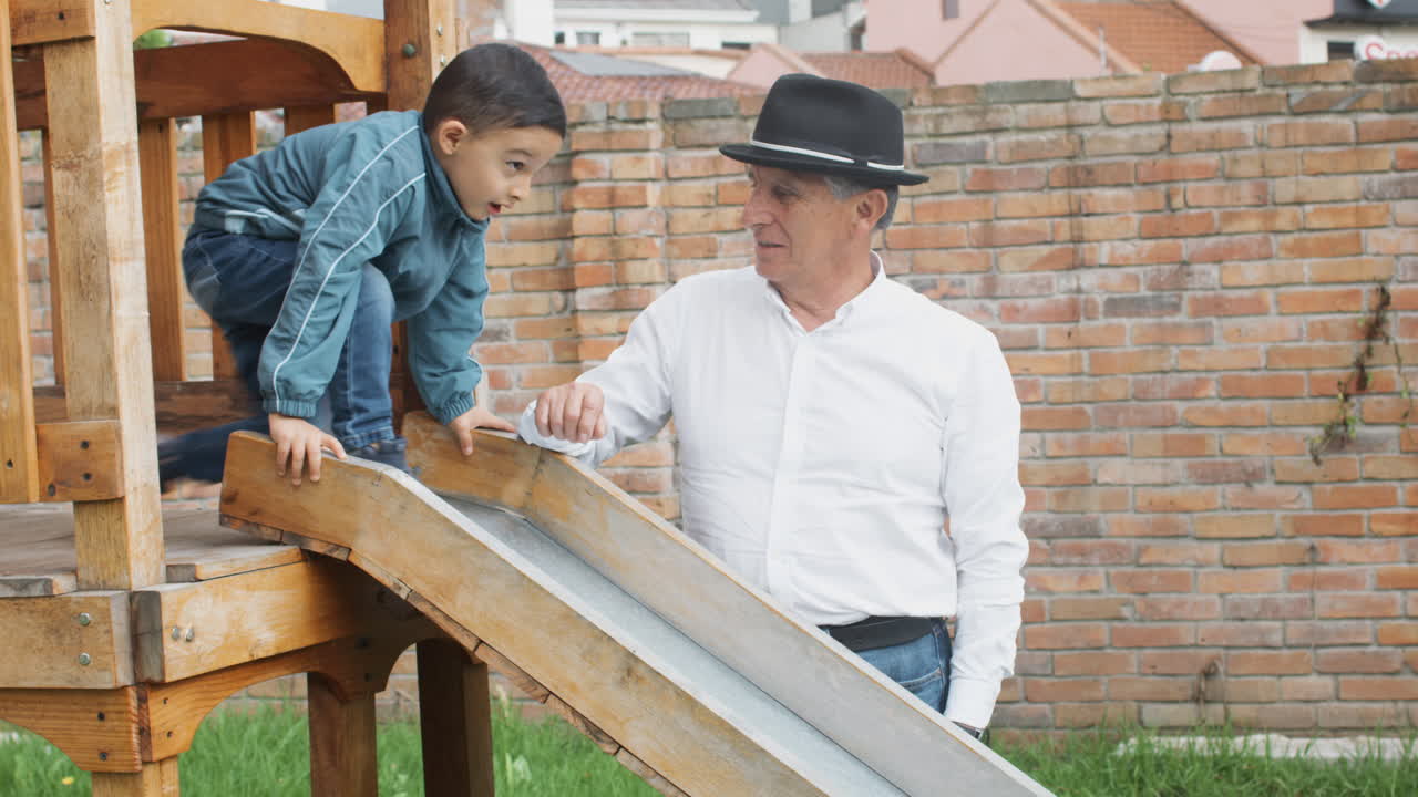 A tender moment between grandfather and grandson as they play together in a wooden playhouse with a slide in the backyard.
