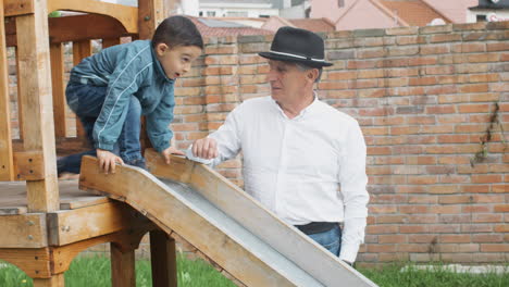a tender moment between grandfather and grandson as they play together in a wooden playhouse with a slide in the backyard.