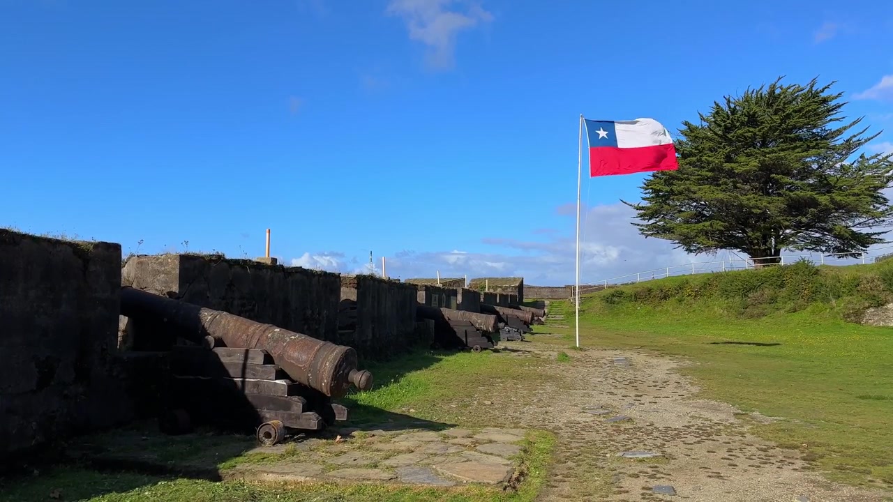 Ampia inquadratura statica della bandiera cilena che sventola vicino a cannoni storici presso il Castello di San Sebastian de la Cruz a Corral.