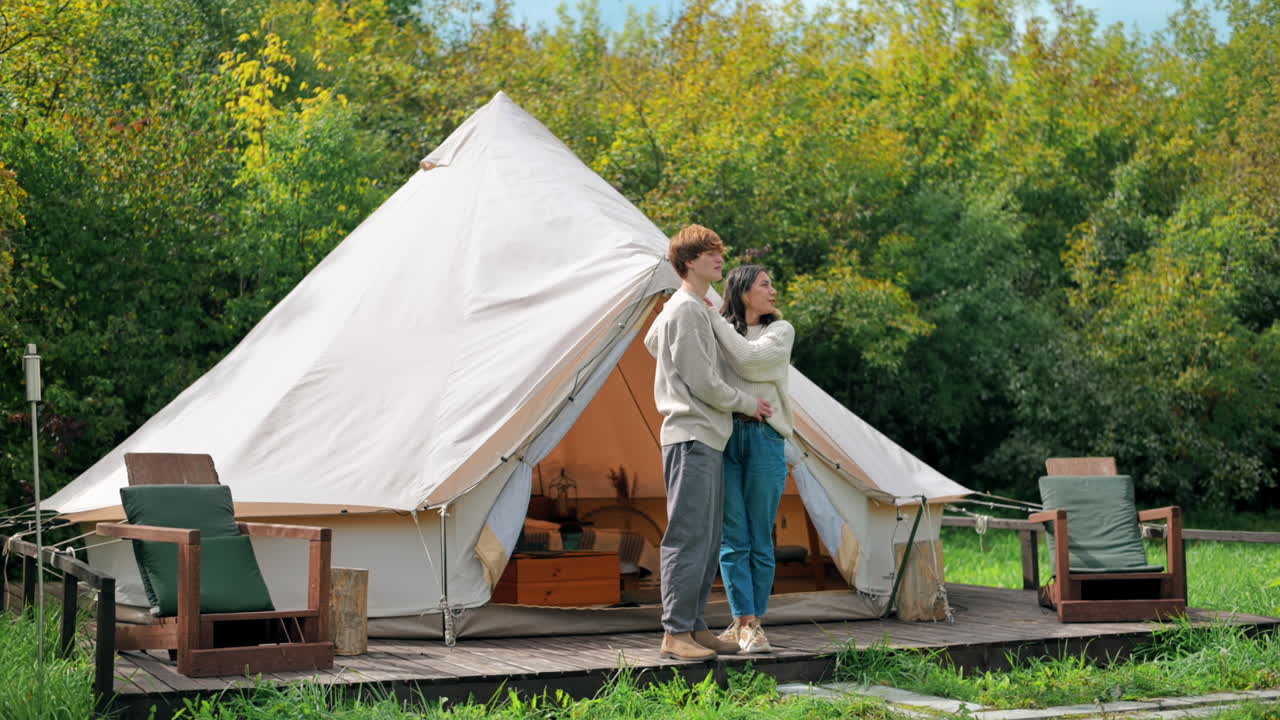 Et par, der omfavner hinanden og snakker på en teltterrasse ved glamping. Frodig skov omkring.