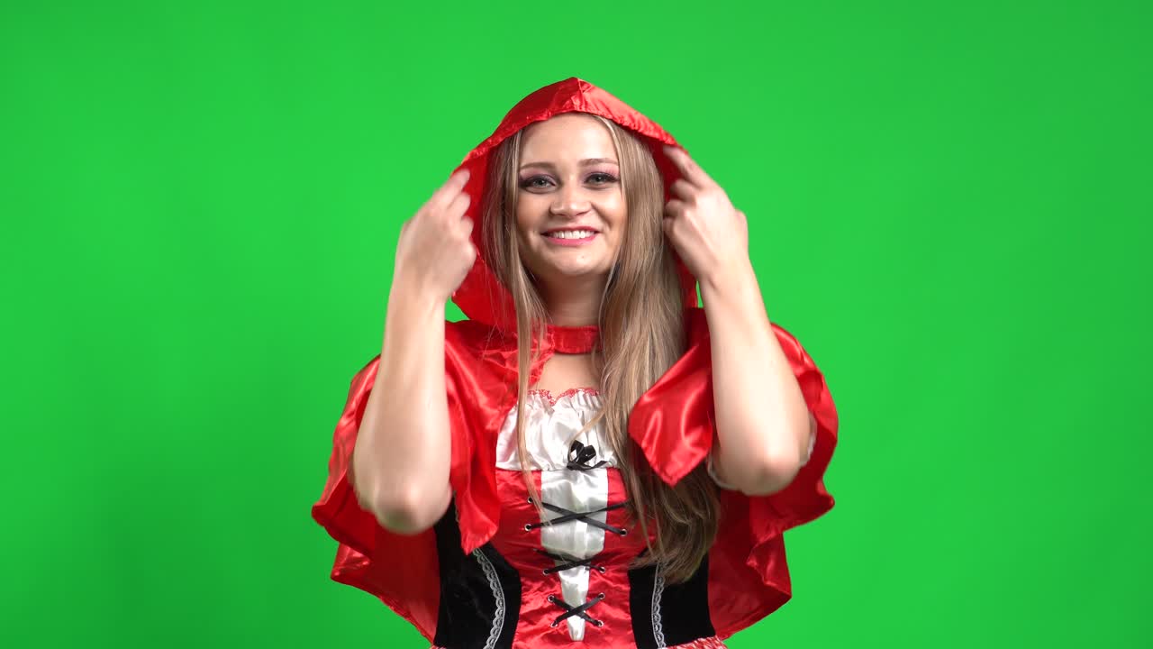 una chica hermosa, vestida con un disfraz de caperucita roja, frente a una pantalla verde, sonriendo mientras mira a la cámara y cubriendo su cabeza con la capucha