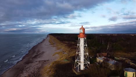 aerial view of white painted steel lighthouse located in pape, latvia at baltic sea coastline in cloudy day, wide angle ascending altitude establishing drone shot moving forward