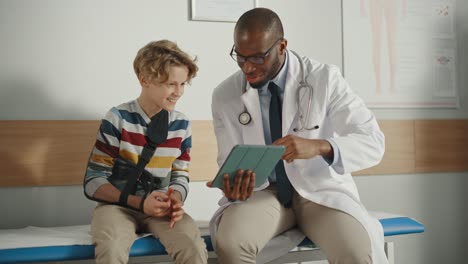 friendly african american family doctor talking with a young boy with arm brace and showing test results on tablet. happy medical care physician in a hospital is reassuring the boy with broken arm.