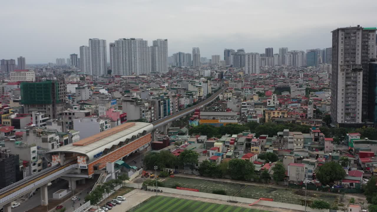 Aerial drone footage of Hanoi Metro elevated railway in Vietnam, showing train station, city buildings, sports field, and high-rise skyline under overcast sky in crowded urban setting