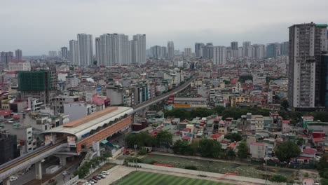 aerial drone footage of hanoi metro elevated railway in vietnam, showing train station, city buildings, sports field, and high-rise skyline under overcast sky in crowded urban setting
