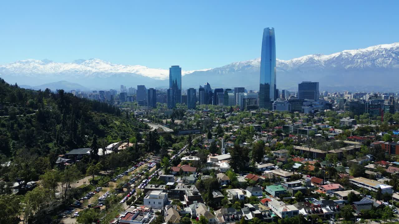 Drone aerial forward approach over residential hills and treetops toward the Costanera skyscrapers and snowcapped Andes in clear daytime light. Santiago, Chile