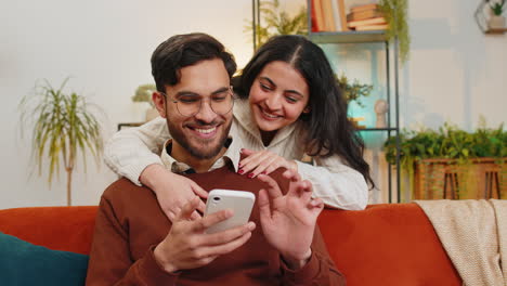 Mujer-India-Sonriente-Hablando-Con-Su-Novio-Deslizando-El-Teléfono-Inteligente-Sentada-En-El-Sofá-En-Casa,-Pareja-Familiar