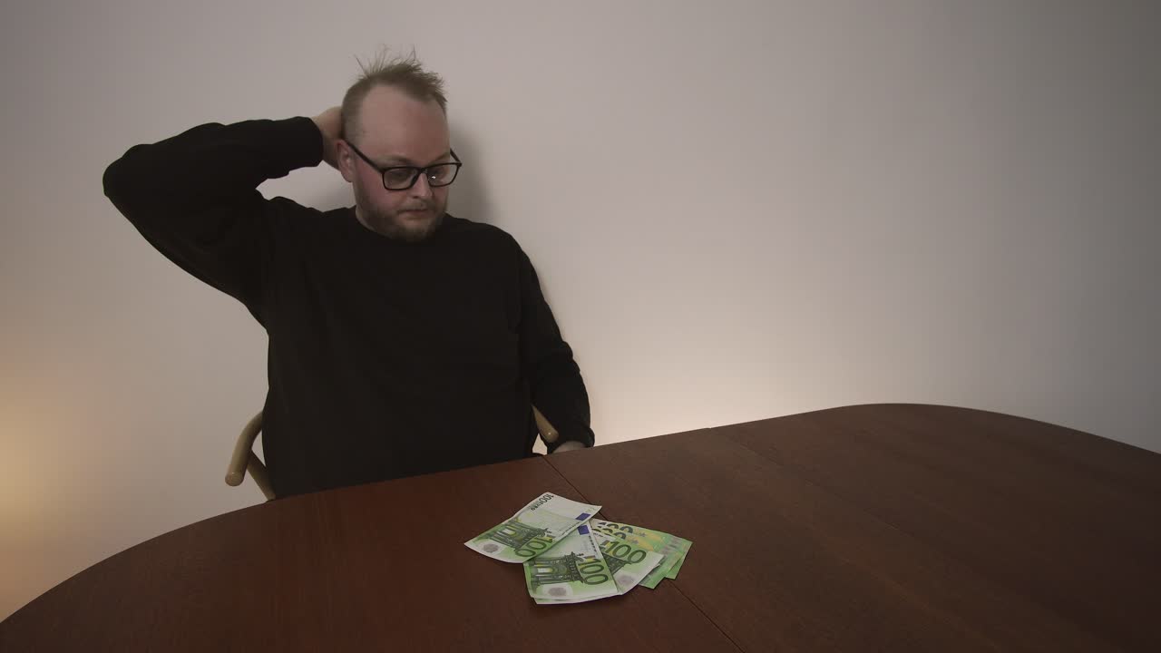 Homem segura a cabeça e balança levemente à mesa com notas de euro. Conceito de estresse financeiro, ansiedade, dívida e pressão orçamentária doméstica.