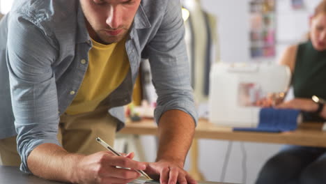 male fashion designer drawing design in foreground as female colleague works on sewing machine