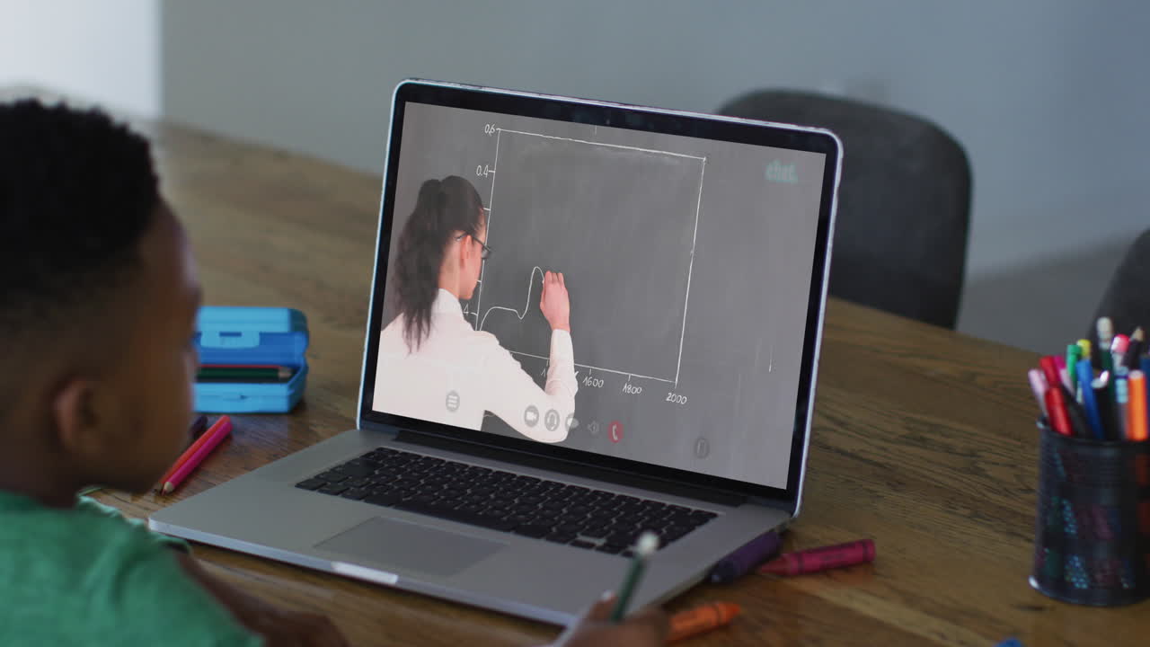 African american boy doing homework while having a video call with female teacher on laptop at home