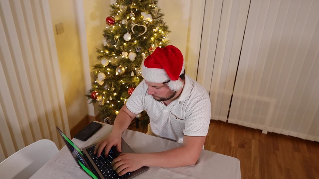 Man in festive Santa hat surfing working from home on a laptop computer online at Christmas with a decorated Christmas tree