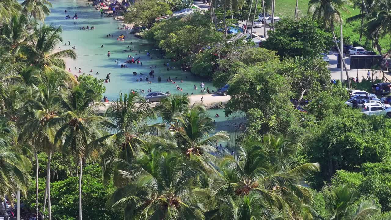 People relax and cool off in a hot day at the Arroyo Salado river resort in the Dominican Republic. Pan left over palm tree canopy