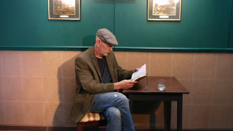 a lonely man wearing a flat cap is seated at a pub table, intently reading a book. a glass sits on the table, while framed artwork hangs on the wall behind him, creating a quiet, focused atmosphere