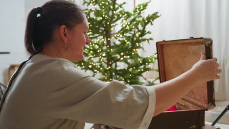 woman opening vintage box filled with colorful ornaments beside decorated christmas tree, joyfully picking up two festive items while smiling warmly in cozy holiday-themed living room