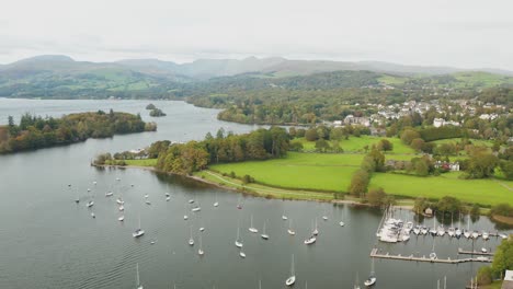 aerial shot from a drone of lake windermere in the foreground there are fields and the small town of bowness in the background you can see the mountains and hills of the lake district