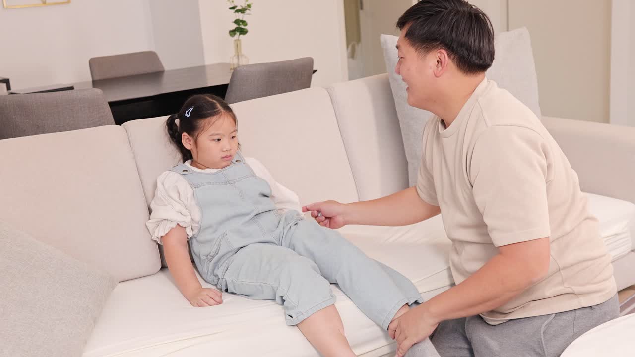 A father tends to his young daughter on a sofa, providing comfort and care in a softly lit living room