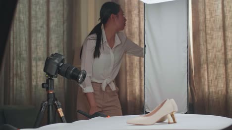fotógrafa asiática ajustando el equipo de iluminación para obtener una buena iluminación en los zapatos de las mujeres mientras toma fotos de los productos en el estudio doméstico