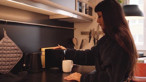 woman in comfy robe in kitchen uses machine to make cup of coffee, profile view