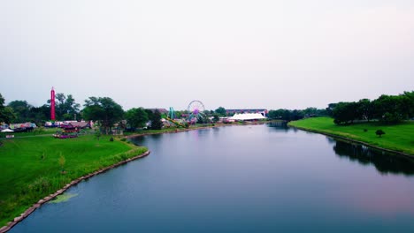 above-Little-Bear-Lake-towards-Carnival-in-Vernon-Hills,-Illinois,-USA-Century-Arboretum-Park---Ferris-Wheel--Carousell-and-tents,-Aerial-Sunset