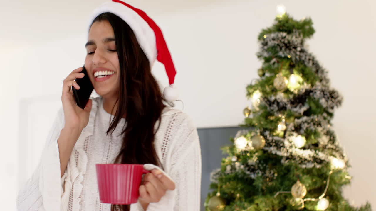 Smiling woman in Santa hat talking on phone, holding mug by Christmas tree
