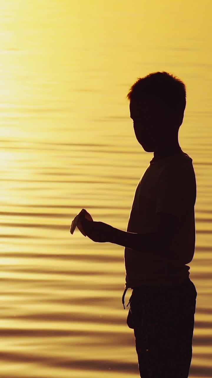 little boy holds a paper airplane in his hand at sunset. A child raised his hand up to sky and plays with origami in the evening in the street. Vertical video