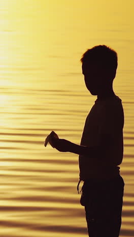 little boy holds a paper airplane in his hand at sunset. a child raised his hand up to sky and plays with origami in the evening in the street. vertical video